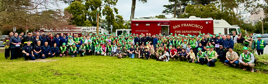 A diverse group of over 150 NERTs and S F F D Staff pose for the official drill photo, with many smiling faces. Behind the NERTs is the S F F D mobile command van.