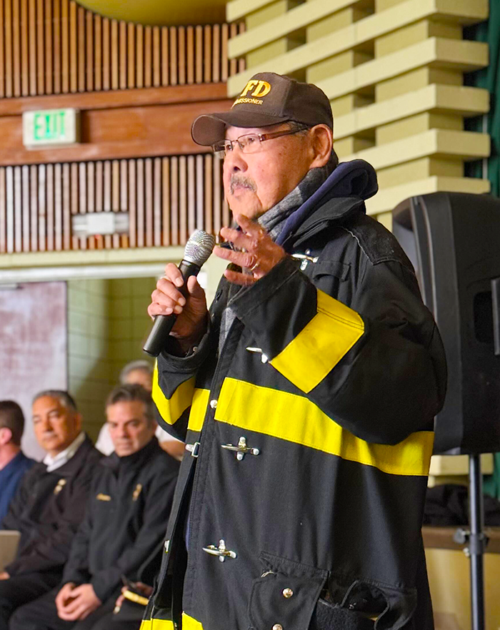 S F F D Commisioner Nakajo holds a microphone, wearing a firefighter coat, speaks to the audience.