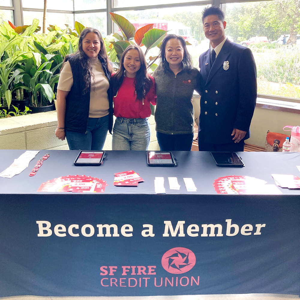 Four people standing shoulder to shoulder, with big smiles. The people are standing infront of a table, with a sign that reads: Become A Member SF Fire Credit Union.